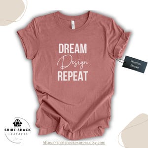 Puede incluir: Camiseta color malva jaspeado con las palabras "DREAM Design REPEAT" en blanco. La camiseta tiene cuello redondo y mangas cortas. Una etiqueta negra dice "Heather Mauve". El logotipo de Shirt Shack Express es visible.