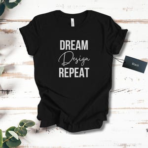 Puede incluir: Camiseta negra con las palabras "DREAM Design REPEAT" en letras blancas en bloque y cursiva. Una etiqueta negra está adherida a la camiseta. La camiseta se muestra sobre una superficie de madera clara.