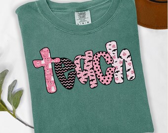 May include: A teal Comfort Colors t-shirt with the word "teach" in a pink and black design. The letters are decorated with patterns including ghosts, bats, polka dots, and zigzags. The shirt is folded and lying flat.