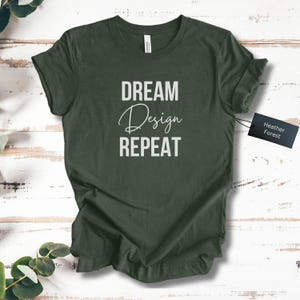 Puede incluir: Una camiseta verde oscuro con las palabras "DREAM Design REPEAT" impresas en blanco. La palabra "Design" está en cursiva. Una etiqueta negra que dice "Heather Forest" está adjunta a la camiseta.