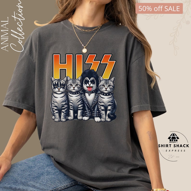 以下が含まれることがあります： KISSのバンドメンバーに扮した4匹の猫のグラフィックがプリントされたグレーのTシャツ。グラフィックには、バンド名「KISS」がスタイリッシュなフォントで、黄色とオレンジのグラデーションで表現されています。「HISS」という文字は、バンド名の真上に、同様のフォントで書かれています。
