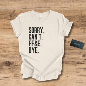 Op de afbeelding: Crèmekleurig T-shirt met de zwarte tekst "SORRY. CAN'T. FF&E. BYE.". Een zwart label met de tekst "Natural" is aan het shirt bevestigd. Het shirt ligt plat op een houten oppervlak.