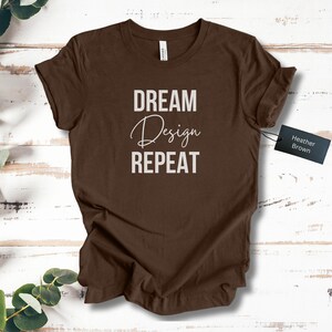 Puede incluir: Una camiseta marrón con las palabras "DREAM Design REPEAT" impresas en blanco. La palabra "Design" está en una fuente cursiva. Una etiqueta negra con "Heather Brown" está sujeta a la camiseta. La camiseta está sobre una superficie de madera.