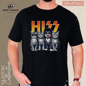 以下が含まれることがあります： KISSのバンドとして扮した4匹の猫のグラフィックが描かれた黒いTシャツ。猫は黒い革のジャケットを着ており、メイクをしており、その上にバンドのロゴ「KISS」があります。