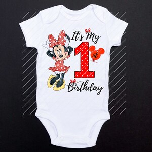 Puede incluir: Un body blanco con un personaje de Minnie Mouse rojo y negro, un n&uacute;mero uno rojo con lunares blancos y el texto "It's My 1st Birthday".