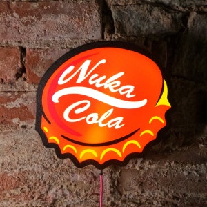 Op de afbeelding: Een neonbord in de vorm van een flesdop, met de woorden "Nuka Cola" in wit op een oranje achtergrond. Het bord heeft een zwarte rand en gele accenten, met een roze stroomkabel.