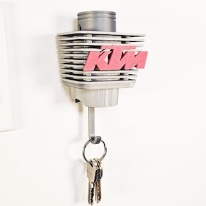 Puede incluir: Un llavero KTM gris y rojo montado en una pared blanca. El llavero tiene forma de cilindro de motor con el logotipo KTM en rojo. Un gancho metálico sostiene un llavero con varias llaves.