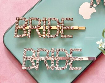 May include: Four rhinestone hair pins spelling "MRS" and "BRIDE" in a script font. The pins are gold-toned and set with clear rhinestones. The pins are displayed on a light blue surface.