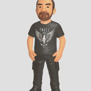 Könnte beinhalten: Eine schwarz-weiße Figur eines Menschen, der ein schwarzes T-Shirt mit einem weißen Adler-Motiv und dem Text "ENGEL 1966" trägt. Die Figur trägt eine schwarze Hose und schwarze Schuhe.
