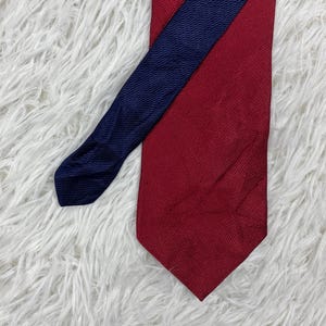 May include: A necktie featuring a deep red main body with a textured, woven fabric surface. A narrower, dark navy blue section with a similar fine, textured weave pattern is layered diagonally across the front of the red tie, creating a contrast.