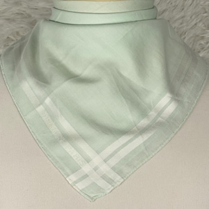 May include: A light sage green square scarf featuring a subtle tonal plaid pattern with white stripes along the edges. The fabric has a smooth texture and is folded into a triangular shape, displaying a clean, uniform color throughout the design.