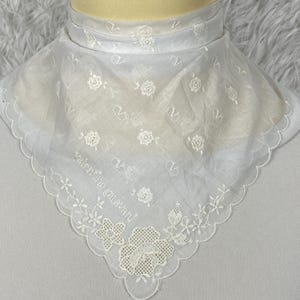 May include: A white sheer triangular scarf featuring delicate white floral embroidery and the Valentino Garavani logo pattern throughout. The edges are finished with a scalloped border and a larger floral embroidery detail at the bottom point.