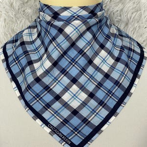 Puede incluir: Un pañuelo a cuadros azul, blanco y marino con el texto "CK CALVIN KLEIN" en la parte inferior. El pañuelo se muestra en una cabeza de maniquí. El patrón de cuadros es un diseño clásico.