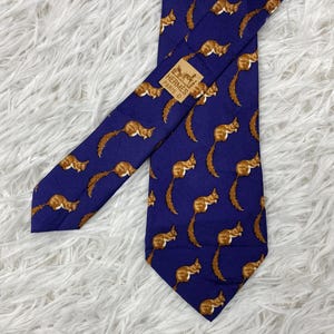 May include: A deep purple necktie featuring a repeating pattern of brown squirrels. The tie includes a rectangular fabric label stitched near the top center that reads HERMÈS PARIS. The design consists of multiple squirrels in various poses.