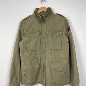May include: Olive green military-style jacket with multiple front pockets and a stand-up collar. Features an American flag patch on the upper sleeve. The jacket is hanging on a wooden hanger against a white background.