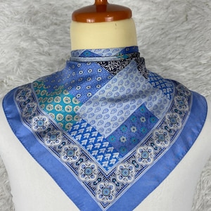 May include: A blue silk scarf with a patchwork design. The scarf features various shades of blue, with intricate patterns including paisley, floral, and geometric shapes. The scarf is displayed on a mannequin.