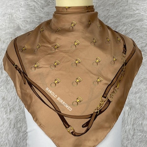 Puede incluir: Un pañuelo cuadrado de seda color beige con un estampado repetido de herrajes ecuestres dorados. El pañuelo tiene un borde marrón con un diseño de cinturón y cadena. El texto "MANOLO BORROMEO" está impreso en el pañuelo.