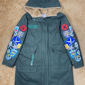 Könnte beinhalten: Olivgrüner Parka mit Kapuze, gefüttert mit Kunstfell. Die Jacke hat mehrere gestickte Aufnäher auf den Ärmeln, darunter die Nummer "093" und militärische Designs. Sie hat zwei Vordertaschen und einen grünen Riemen.