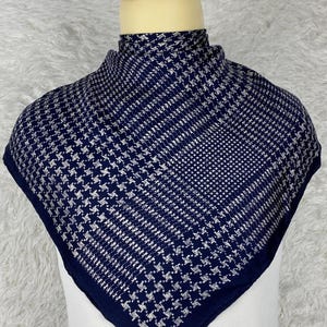 May include: A navy blue and white houndstooth patterned scarf. The square scarf is displayed on a mannequin head and neck. The scarf has a dark blue border and a repeating pattern of white and navy blue shapes.
