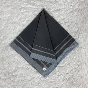 Puede incluir: Un pañuelo gris oscuro doblado con rayas azules y grises y una pequeña etiqueta blanca. El pañuelo tiene forma cuadrada y descansa sobre una superficie blanca y esponjosa.