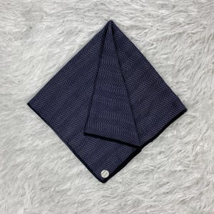 May include: A navy blue handkerchief with a white zigzag pattern and a black border. The handkerchief is folded in a diamond shape and has a small, round tag in the corner.