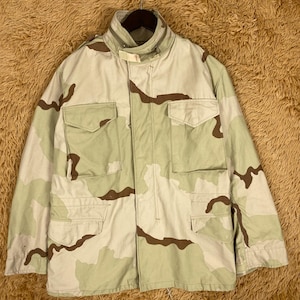 May include: A desert camouflage jacket with multiple front pockets and a high collar. The jacket features a tan, brown, and light green pattern. It is hanging on a hanger.