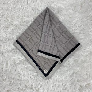 Puede incluir: Un pañuelo gris plegado con un patrón de cuadros y borde negro. El logotipo de Calvin Klein es visible en una pequeña etiqueta. El pañuelo está hecho de una tela suave y está doblado en forma cuadrada.
