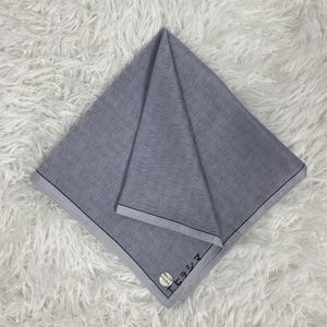 Puede incluir: Un textil cuadrado gris doblado con el logotipo de Calvin Klein repetido en la superficie. Los bordes están recortados con un fino borde negro. Una pequeña etiqueta blanca y negra con texto japonés es visible.