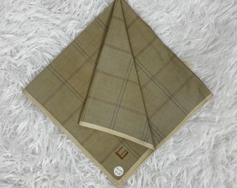 Può includere: Un fazzoletto beige piegato con un motivo a quadri marrone e nero. Il fazzoletto ha un bordo color crema e un piccolo logo nell'angolo. Il logo include il testo "dunhill" e un piccolo logo circolare.