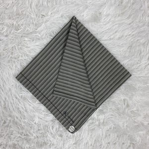 Puede incluir: Una tela plegada a rayas grises y blancas, posiblemente un pañuelo o bufanda, se muestra sobre una superficie blanca y esponjosa. La tela tiene una textura acanalada y una pequeña etiqueta con texto y un logotipo en la esquina.