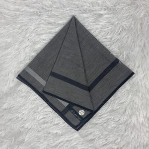 Puede incluir: Un pañuelo gris doblado con rayas azul oscuro y plateadas. El pañuelo tiene una pequeña etiqueta blanca con texto. La tela está doblada en forma de triángulo.