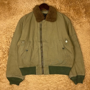 Puede incluir: Chaqueta bomber verde oliva con cuello de piel marrón. La chaqueta tiene cierre de cremallera, dos bolsillos delanteros y puños y dobladillo de canalé verdes. Cuelga de una percha de metal.