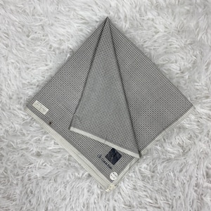 Puede incluir: Un pañuelo a cuadros grises y blancos doblado. La tela está doblada en forma de triángulo, con un borde blanco visible. El pañuelo descansa sobre una superficie blanca y esponjosa. Una etiqueta de marca es visible.