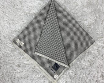 May include: A folded gray and white checkered handkerchief. The fabric is folded into a triangle shape, with the edges showing a white border. The handkerchief is lying on a white, fluffy surface. The brand name is visible on a small tag.