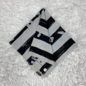 May include: Folded square scarf with a gray background and bold black brushstroke stripes. The scarf has the text "Issey Miyake" printed on it, along with a small circular tag.