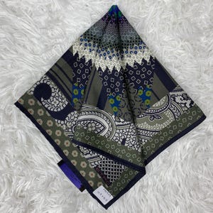 May include: A patterned square scarf in green, blue, and white. The design features geometric shapes, paisley motifs, and floral details. The scarf has a label with text and measurements of 163 x 163 cm.