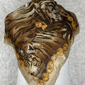 Könnte beinhalten: Ein quadratisches Seidentuch mit Tigerprint in Braun- und Beigetönen. Der Schal hat ein goldfarbenes Kettenbordürendesign. Der Schal ist über einer Schaufensterpuppe drapiert.