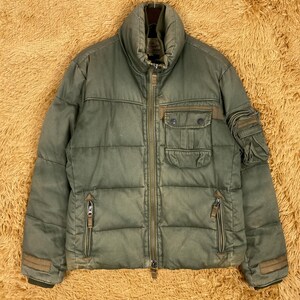 May include: Olive green puffer jacket with a high collar, multiple pockets, and a full-length zipper. The jacket has brown accents on the pockets and sleeves. The jacket is hanging on a metal hanger.