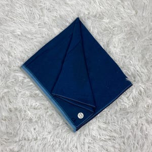 Puede incluir: Una tela azul marino doblada con un borde azul claro. La tela cuadrada parece estar hecha de un material suave. Una pequeña etiqueta blanca con texto es visible en la esquina. La tela se muestra sobre una superficie blanca y esponjosa.