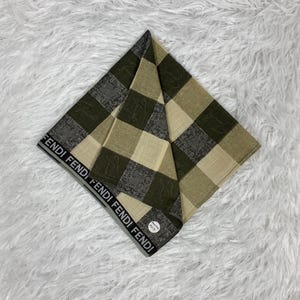 May include: A folded square scarf with a checkered pattern in shades of green, gray, and beige. The border features the brand name "FENDI" in a repeating black and white design. The scarf is displayed on a white, fluffy surface.