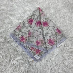 Peut inclure: Un foulard carré à motifs floraux avec des lys roses et des feuilles grises sur fond blanc. Le foulard a une bordure bleu clair. Le foulard est plié en losange. Il y a une étiquette avec du texte.