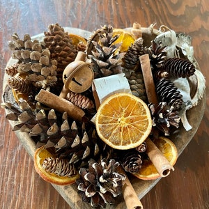 May include: A heart-shaped wooden bowl filled with pine cones, cinnamon sticks, dried orange slices, and a small bell. The bowl is on a wooden table.