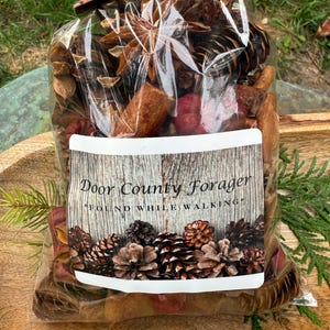 May include: A clear cellophane bag filled with potpourri, including pine cones, star anise, and cinnamon sticks. The label reads "Door County Forager" and "Found While Walking". The bag is tied with twine and a small wooden accent.