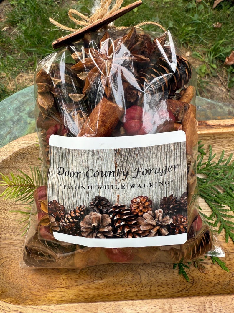 May include: A clear cellophane bag filled with potpourri, including pine cones, star anise, and cinnamon sticks. The label reads "Door County Forager" and "Found While Walking". The bag is tied with twine and a small wooden accent.