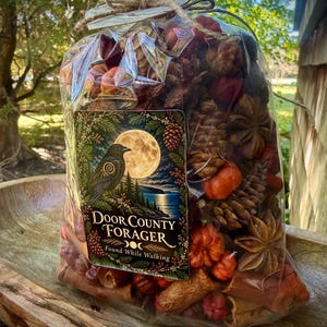 May include: A clear plastic bag filled with dried potpourri, including pinecones, star anise, cinnamon sticks, and small orange gourds. A rectangular label features a black crow, a full moon, and the text Door County Forager Found While Walking.