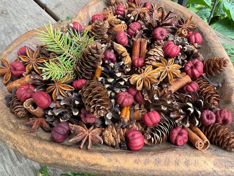 May include: A wooden heart-shaped bowl filled with a decorative arrangement. The arrangement includes pine cones, star anise, cinnamon sticks, small red pumpkins, and sprigs of evergreen. The colors are natural browns, reds, and greens.