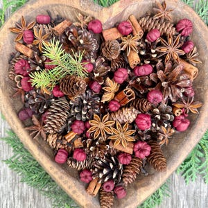 May include: A heart-shaped wooden bowl filled with a decorative arrangement of pine cones, star anise, cinnamon sticks, and small red decorative items. Green sprigs of evergreen are placed around the bowl.