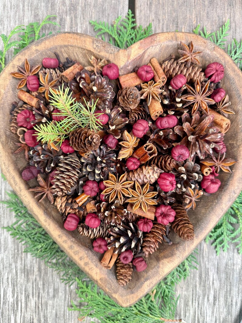 May include: A heart-shaped wooden bowl filled with a decorative arrangement of pine cones, star anise, cinnamon sticks, and small red decorative items. Green sprigs of evergreen are placed around the bowl.