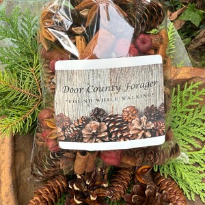 May include: A clear cellophane bag filled with a mix of dried botanicals, including pine cones, star anise, cinnamon sticks, and red berries. The bag has a label that reads "Door County Forager" and "Found While Walking". The bag is tied with a natural twine.
