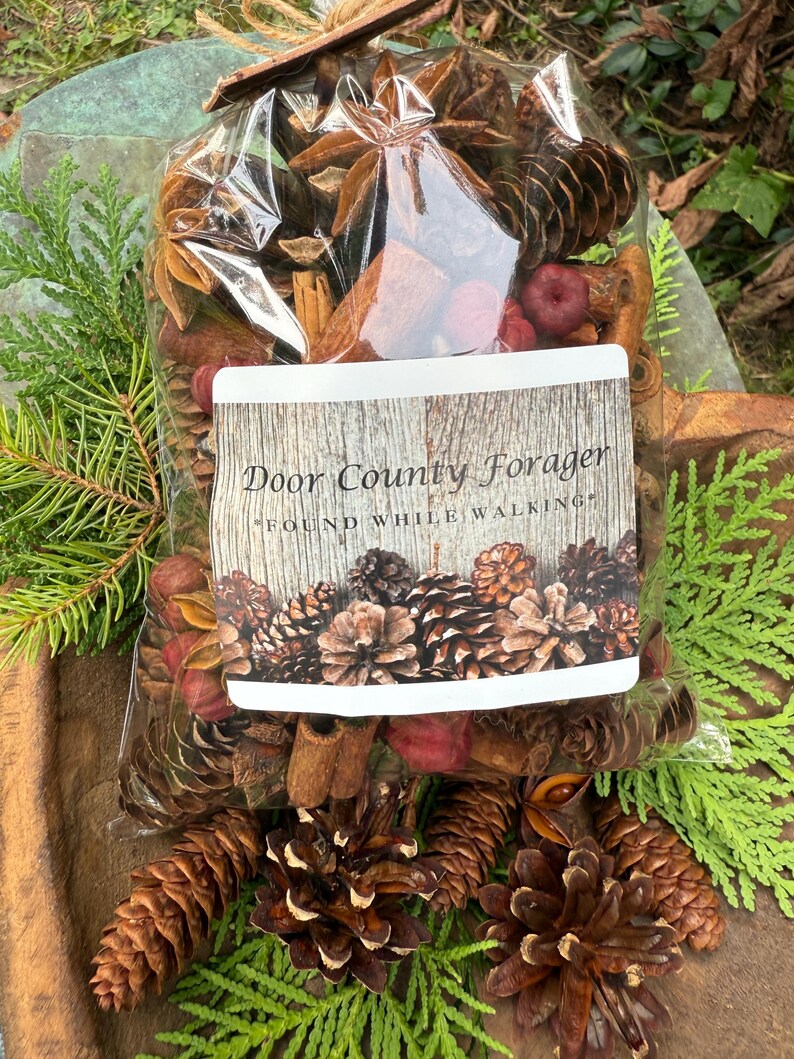 May include: A clear cellophane bag filled with a mix of dried botanicals, including pine cones, star anise, cinnamon sticks, and red berries. The bag has a label that reads "Door County Forager" and "Found While Walking". The bag is tied with a natural twine.